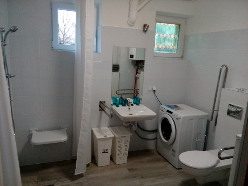 Łazienka w mieszkaniu wspomaganym - sedes, umywalka, kosz na pranie, natrysk prysznicowy z siedziskiem